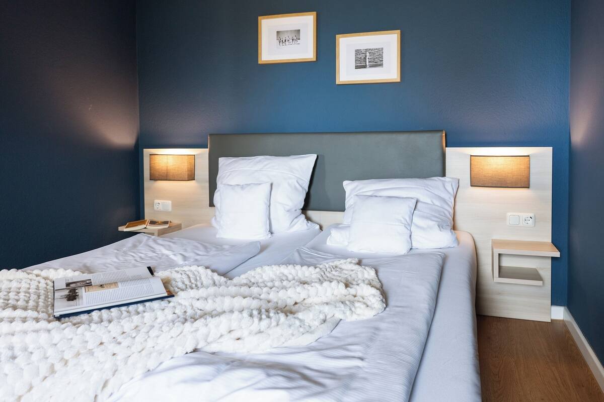 A neatly made bed with white linens, a fluffy white blanket, and a book on the side. There are two bedside tables with lamps, and the walls are painted blue.