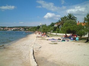 Ein Strand mit badenden und sonnenbächelnden Personen, einem klaren Himmel und Häusern im Hintergrund.