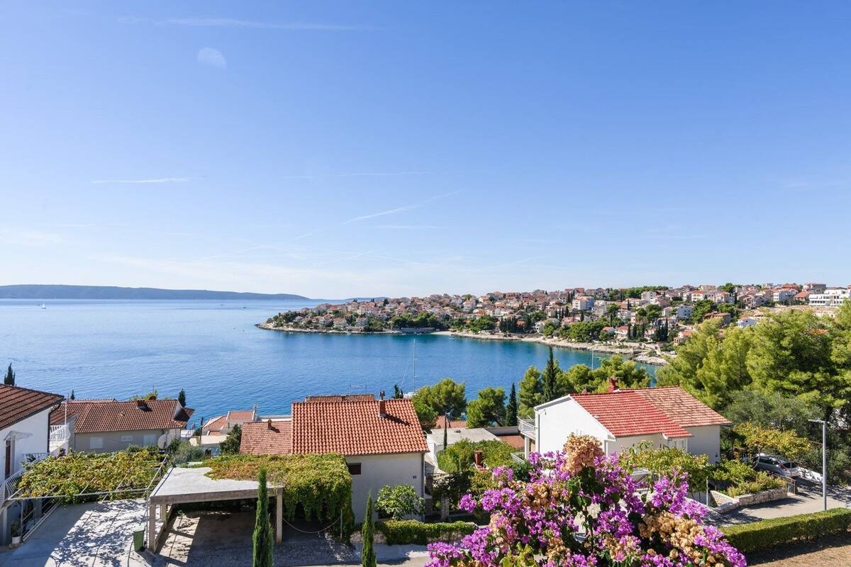 A coastal town with terracotta-roofed houses, a clear blue sea, and a distant view of a larger town across the water.
