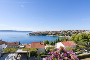 A coastal town with terracotta-roofed houses, a clear blue sea, and a distant view of a larger town across the water.