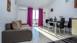 Ein modernes Wohnzimmer mit einer Couch, einem Esstisch mit Stühlen, einem Fernseher und einem Balkon mit violetten Vorhängen.