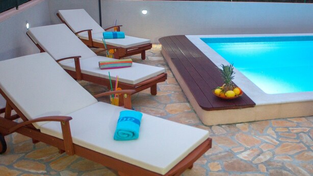 A poolside area with lounge chairs, a small table with a pineapple and fruit, and a stone-tiled floor. - Villa Diana Razanj (Rogoznica)
