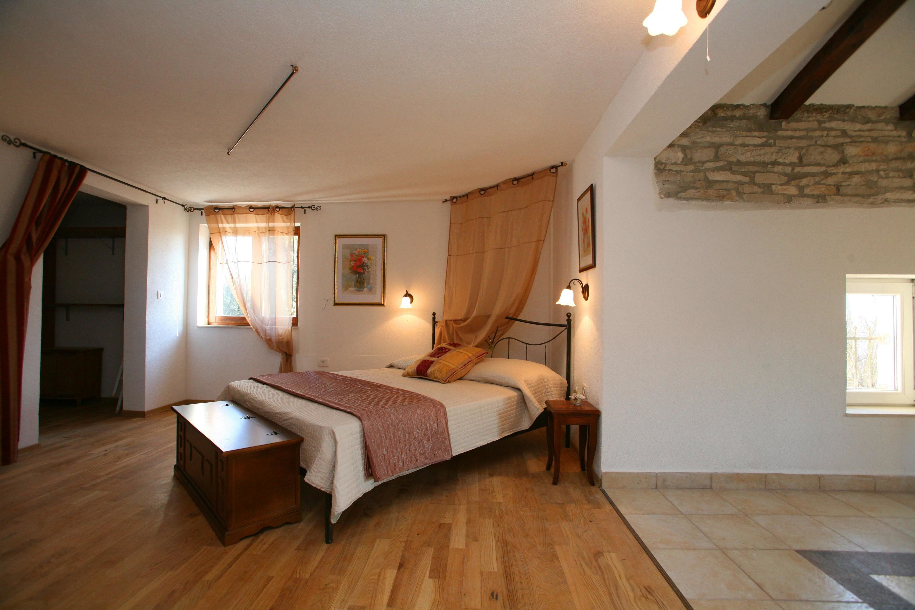 Ein Schlafzimmer mit Holzboden, einem Bett mit Kopfteil, einem Nachttisch, einem Fenster mit Vorhängen und einer Steinwand.