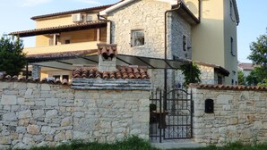 Una casa de piedra de dos pisos con techo de tejas, un muro de piedra con un portón y un pequeño balcón.