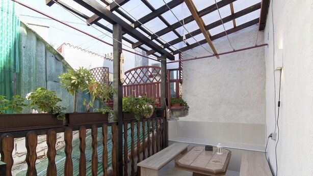 A covered patio with a wooden bench and table, a green fence, and potted plants.