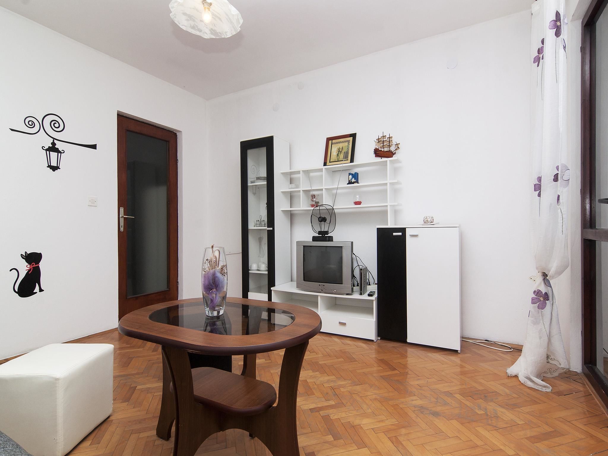 A compact living space with a wooden floor, a glass-top table, a sofa, a TV, and a refrigerator.