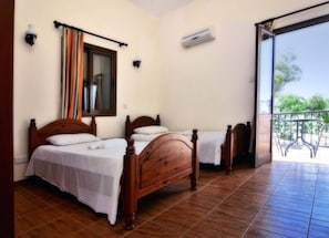 A room with two wooden beds, a window with curtains, an air conditioning unit, and a balcony with a table and chairs.