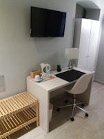 Un espacio de oficina moderno con un escritorio blanco, una silla, un televisor montado en la pared, un armario y un pequeño banco de madera.