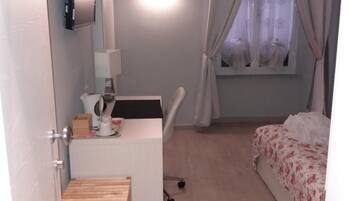 Ein kleines, gut beleuchtetes Schlafzimmer mit Schreibtisch, einem Bett mit floralem Bettbezug, einem Fenster mit transparenten Vorhängen und einer hölzernen Bank.