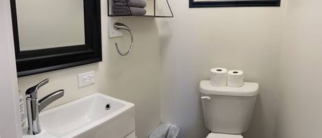 Un baño con un lavamanos blanco, un inodoro, un cubo de basura, un perchero para toallas y un cuadro en la pared.