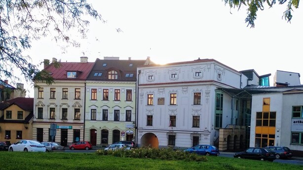 A row of historic buildings with classic architecture, a grassy area in front, and parked cars along the street. - Upper Apart (Cieszyn)