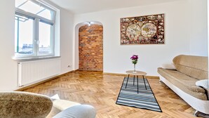 A living room with a herringbone wood floor, a brick wall, a large window, a beige sofa, and a decorative wall piece. - Upper Apart (Cieszyn)