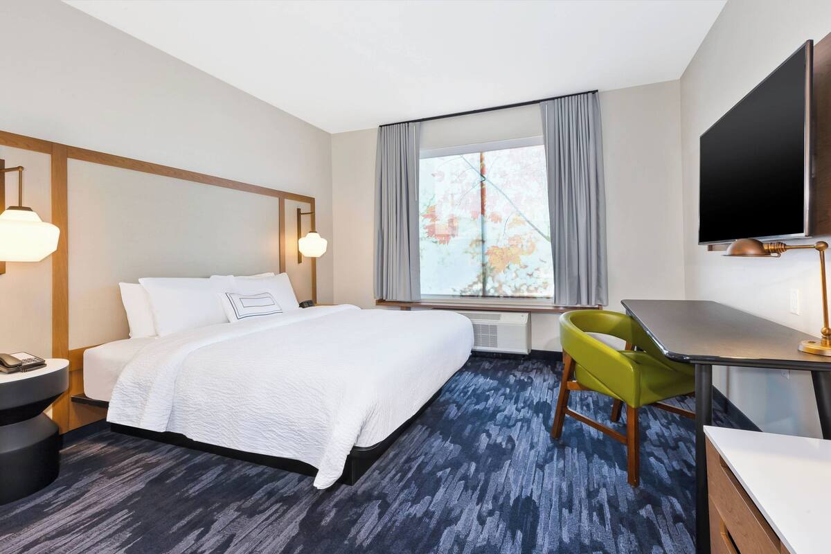 Una habitación de hotel con cama, escritorio, silla, televisor y ventana con cortinas.