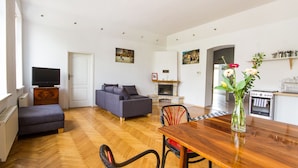 Sala de estar moderna com sofá, mesa de jantar e cadeiras. Há uma lareira, uma televisão e uma área de cozinha visível.