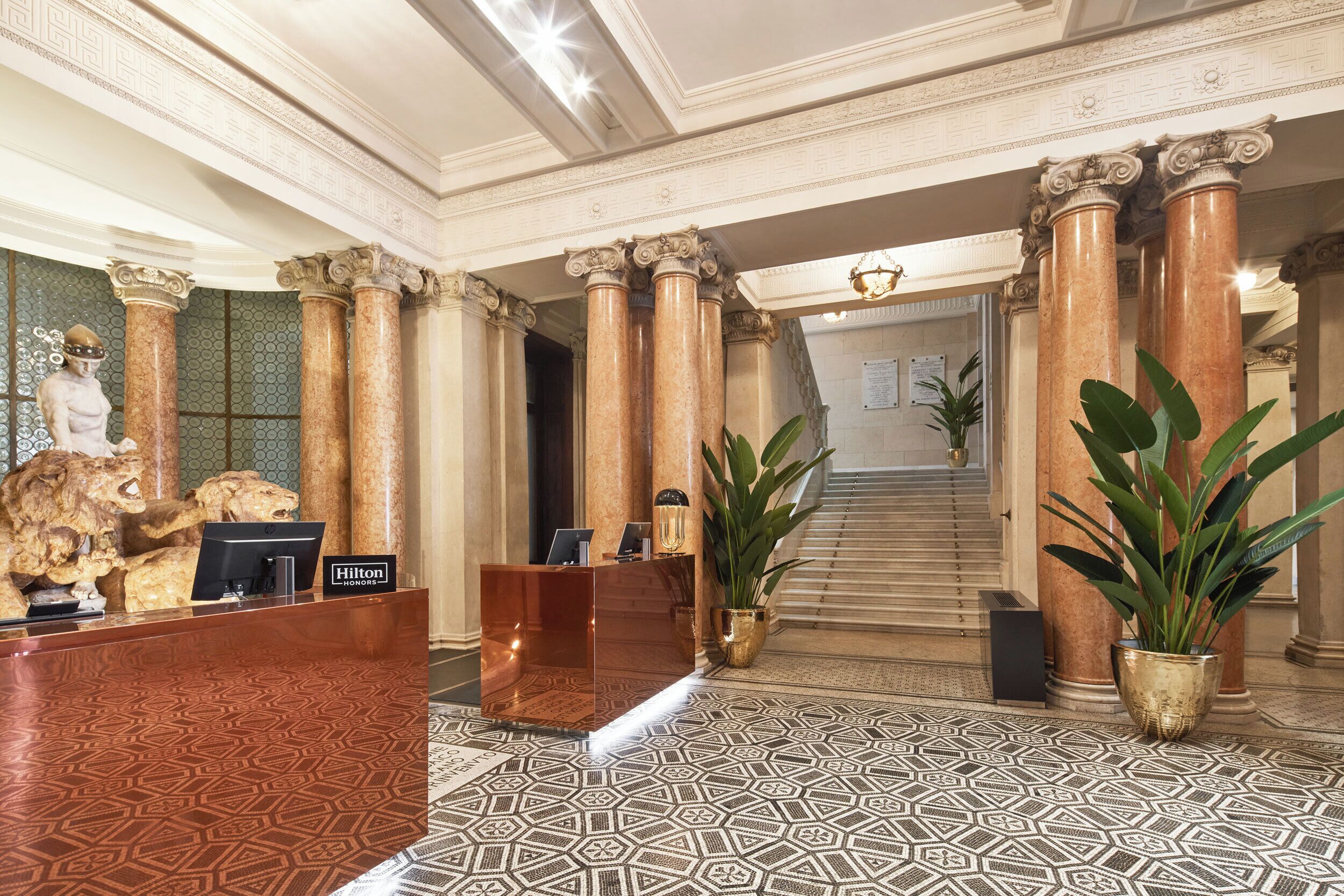 A lobby with a reception desk, marble columns, a statue, and a staircase.