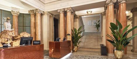 A lobby with a reception desk, marble columns, a statue, and a staircase.