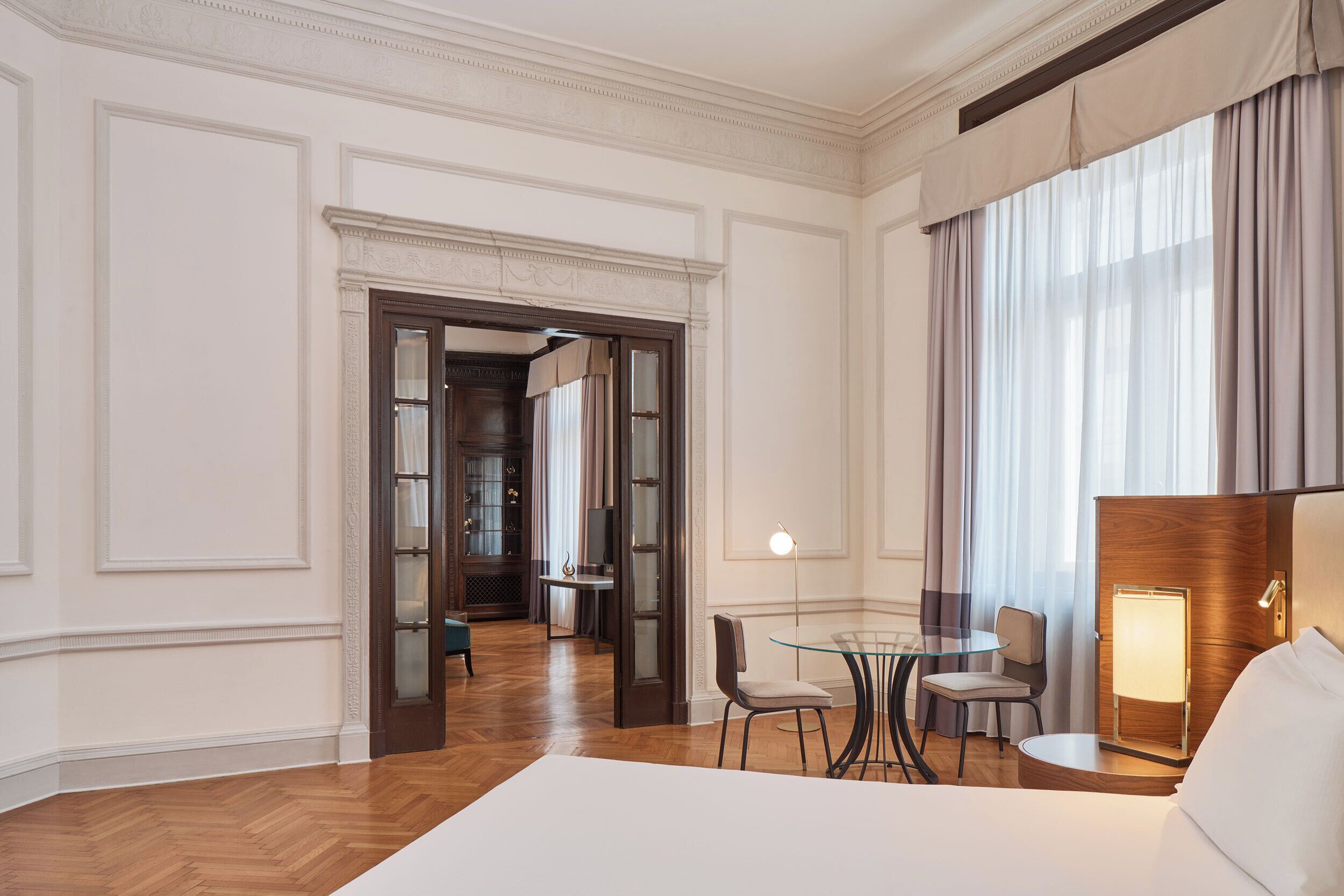 Habitación de hotel con una cama grande, una mesa de vidrio con sillas, un escritorio con lámpara y vistas al pasillo.
