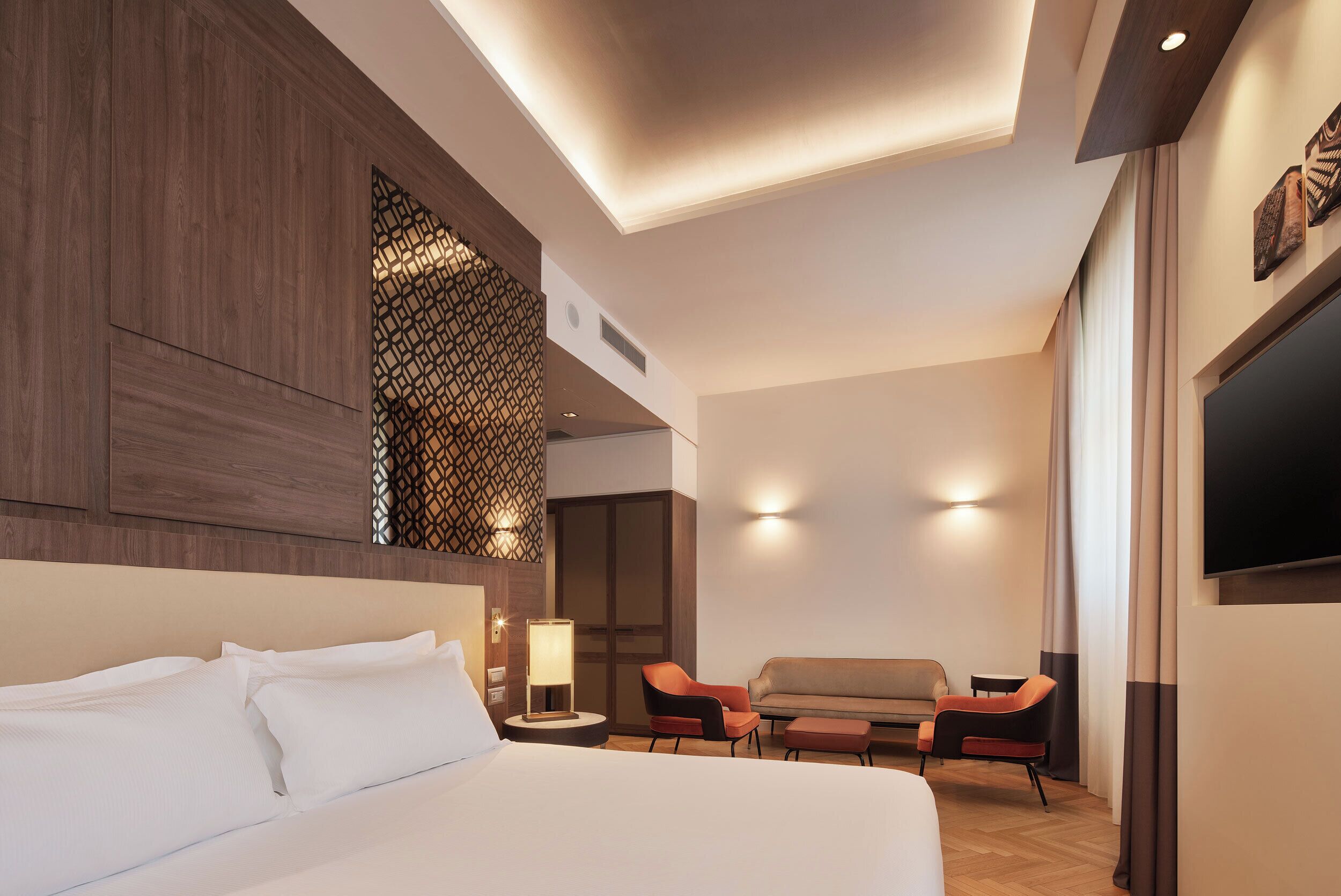 A modern hotel room with a large bed, a seating area with two chairs, a small table, and a TV mounted on the wall.