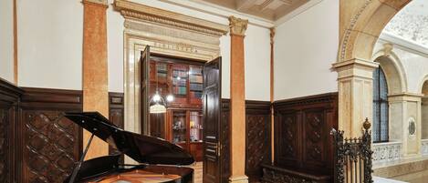A grand piano in a spacious room with ornate wooden paneling, a large bookshelf, and a view of a courtyard through an arched doorway.