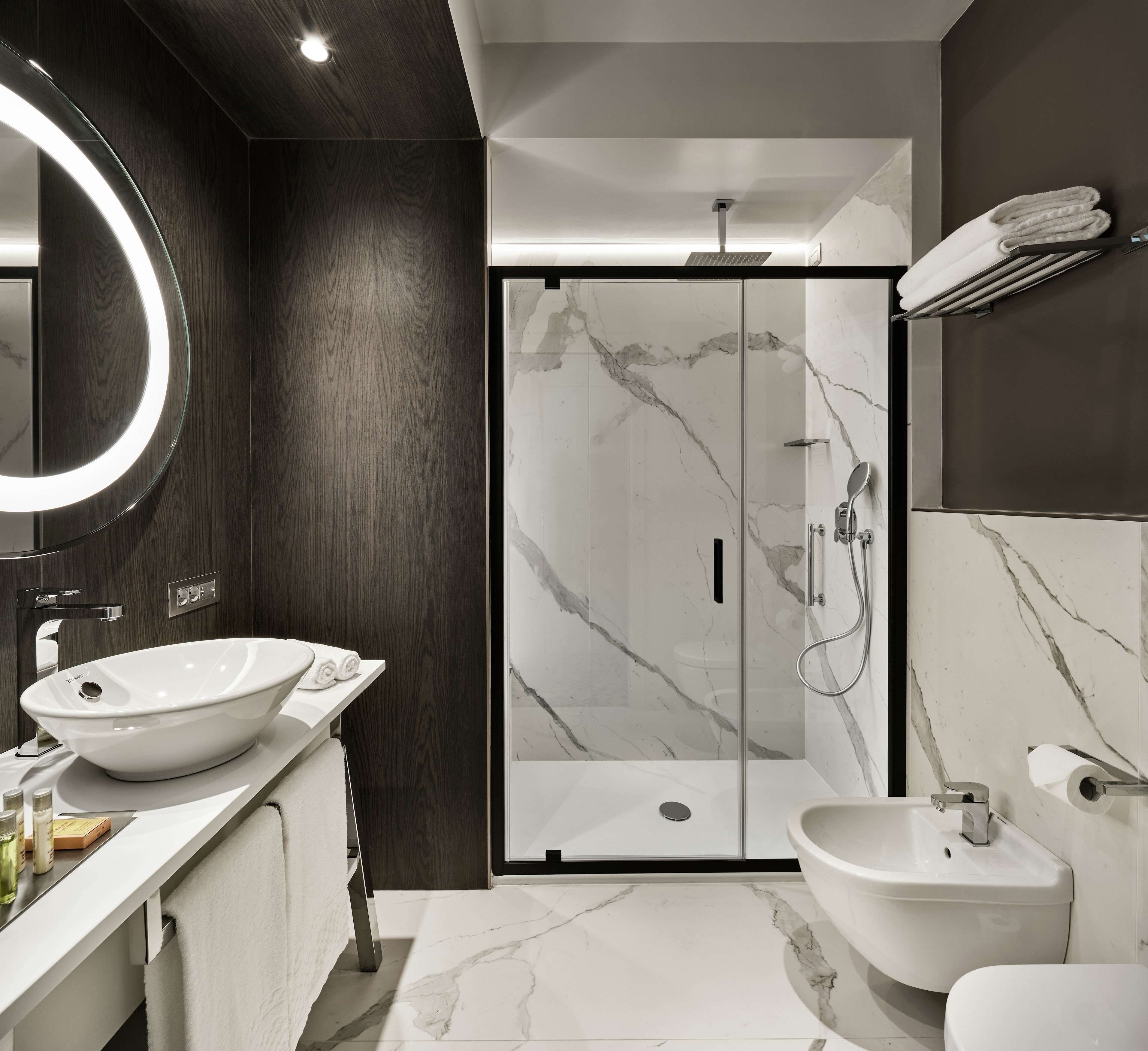 Een moderne badkamer met een douchecabine van glas, een grote ronde spiegel en een witte wastafel.