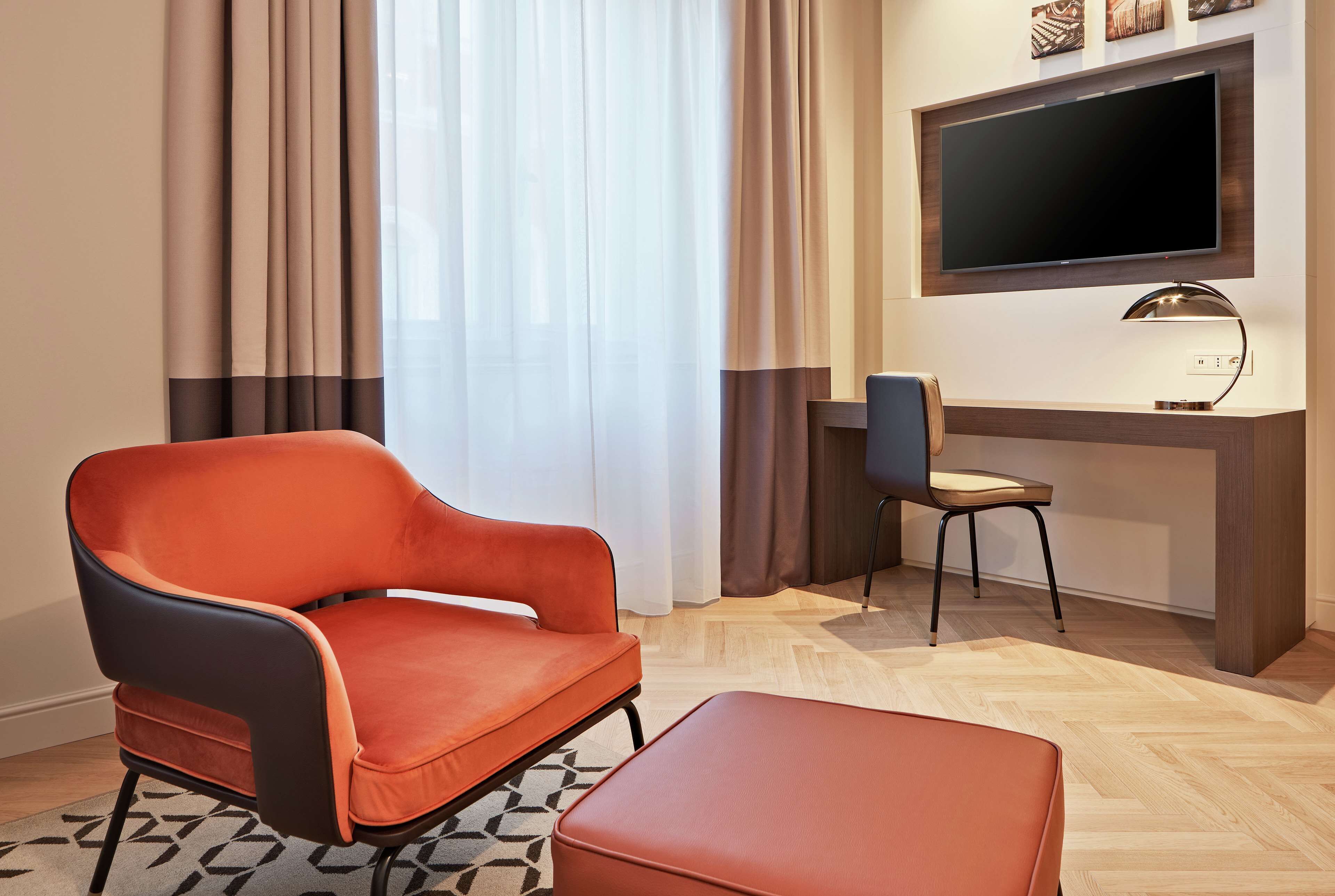 A modern living room with a large flat-screen TV mounted on the wall, a comfortable orange armchair, a matching ottoman, and a desk with a chair.