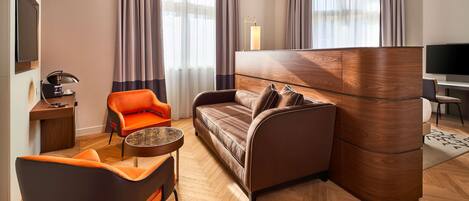 Una habitación de hotel moderna con piso de madera en patrón de espiga, un sofá marrón, un sillón naranja, una mesita redonda pequeña, un televisor y una lámpara.
