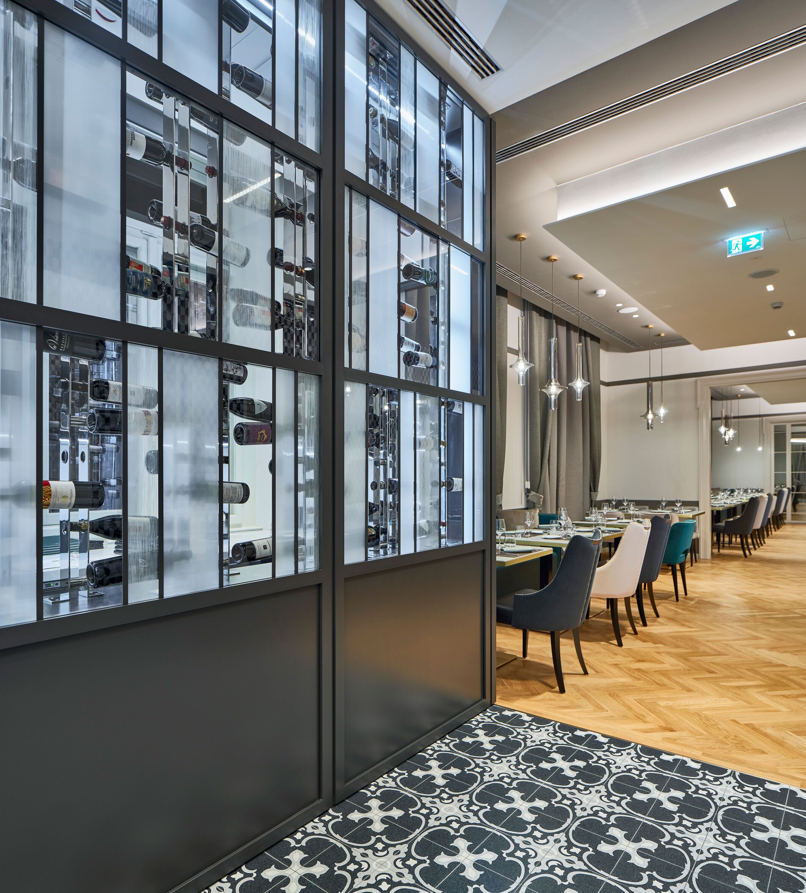 A modern restaurant with a glass wall displaying bottles, wooden flooring, and a long dining area with chairs.