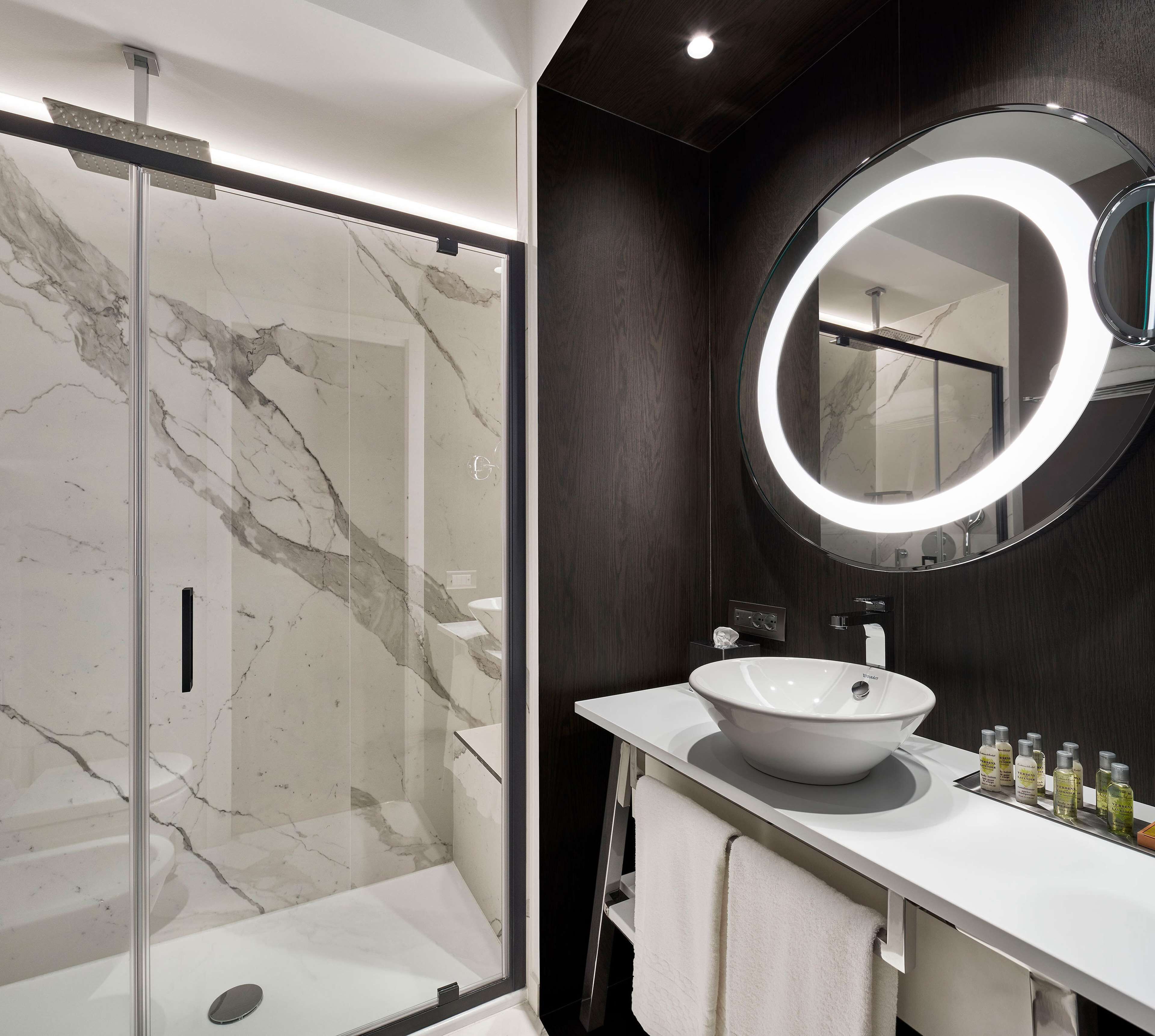 A modern bathroom with a glass shower enclosure, a large round mirror, and a white sink with toiletries.