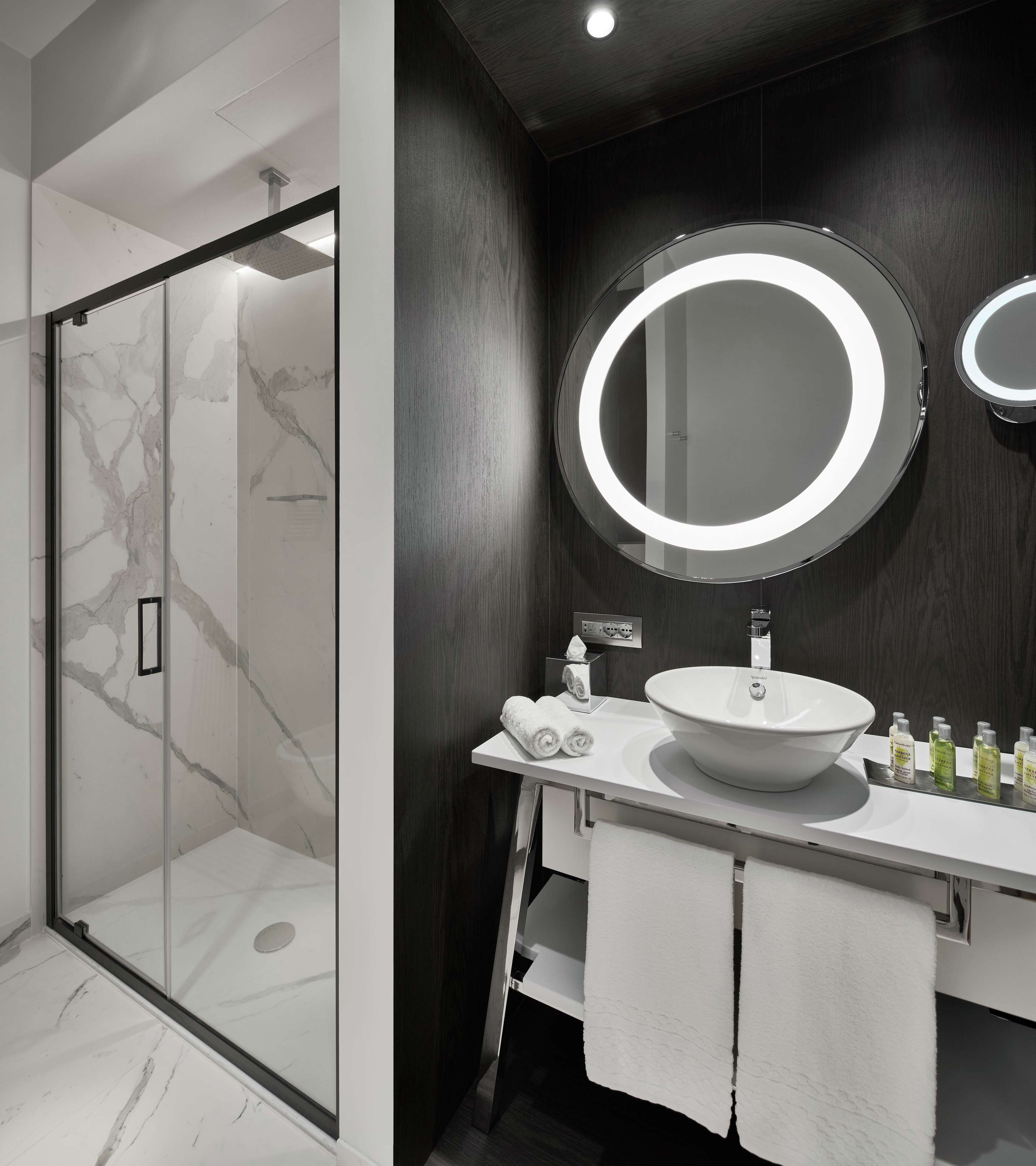 Une salle de bain moderne avec une douche à l’italienne en verre, un meuble-lavabo blanc doté d’un grand miroir rond et un mur revêtu de panneaux en bois foncé.