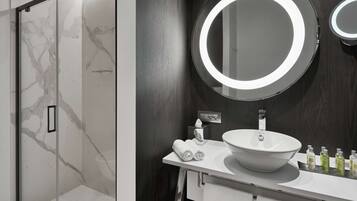 Une salle de bain moderne avec une douche à l’italienne en verre, un meuble-lavabo blanc doté d’un grand miroir rond et un mur revêtu de panneaux en bois foncé.