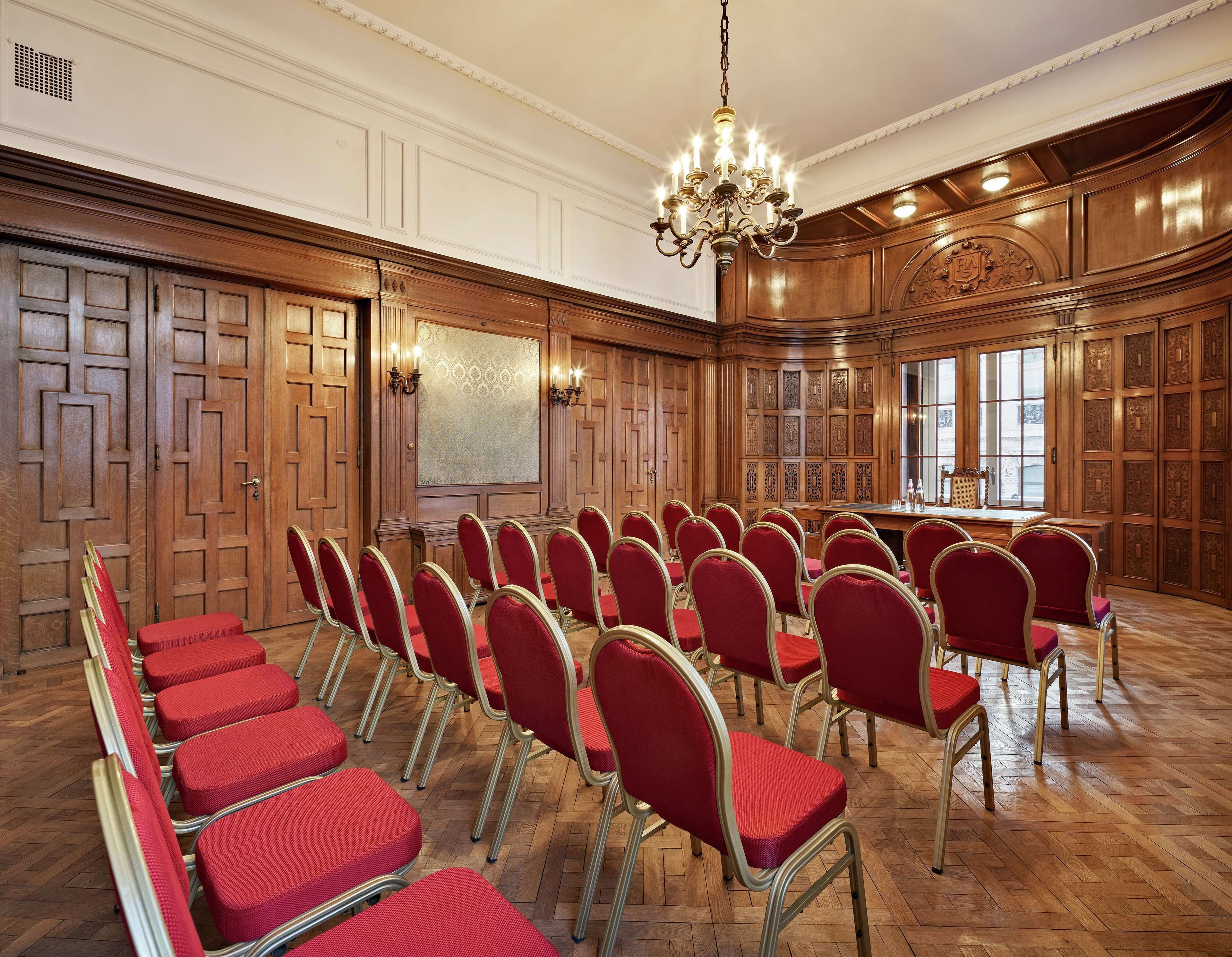 A conference room with wooden paneling, a chandelier, and rows of red chairs.