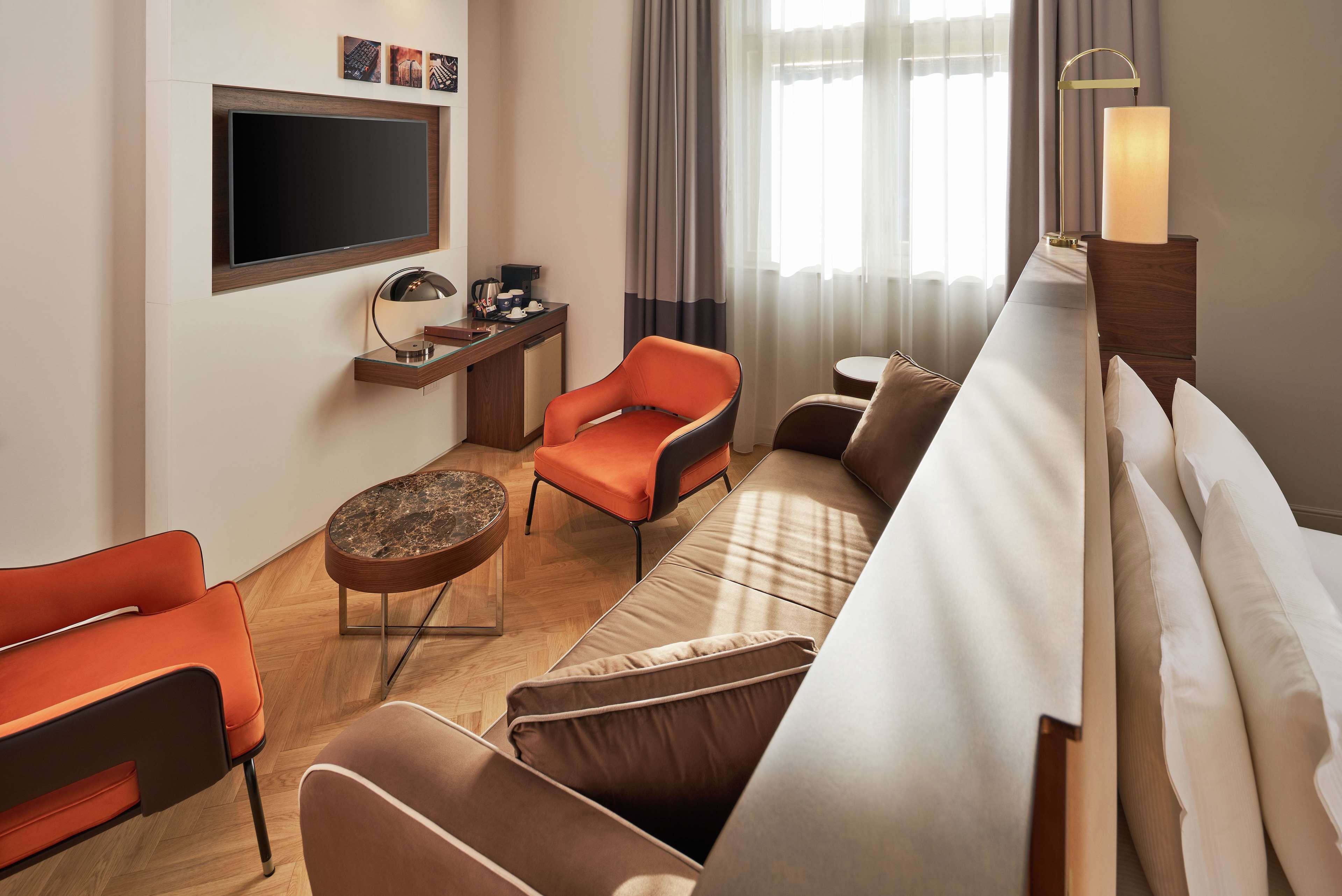Une chambre d’hôtel moderne équipée d’un téléviseur à écran plat fixé au mur, d’un bureau en bois avec une lampe, d’un canapé et d’un fauteuil orange.