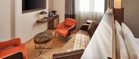 Une chambre d’hôtel moderne équipée d’un téléviseur à écran plat fixé au mur, d’un bureau en bois avec une lampe, d’un canapé et d’un fauteuil orange.