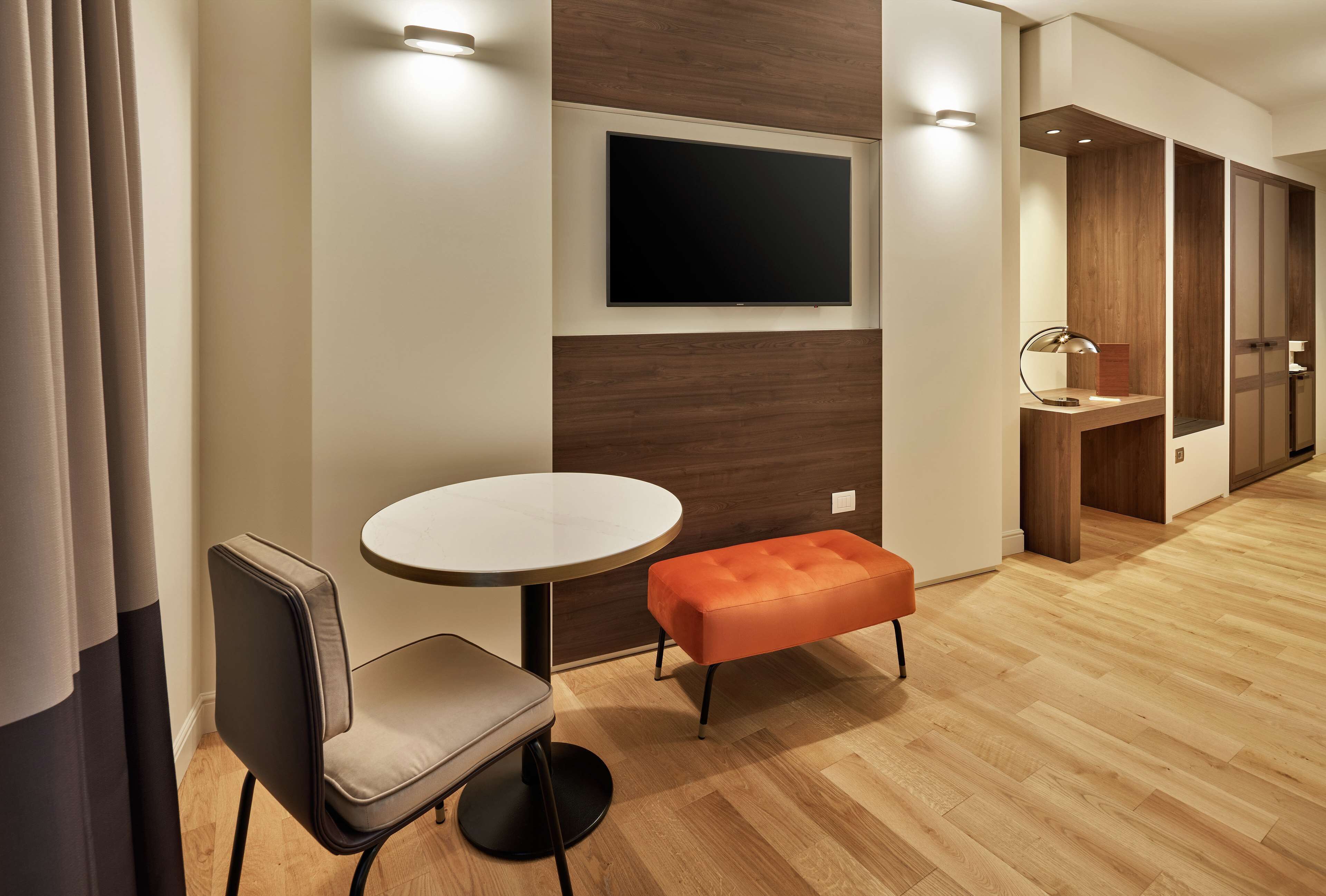 Una habitación de hotel moderna con una mesa redonda, un banco, un televisor de pantalla plana en la pared, un tocador de madera y piso de madera.