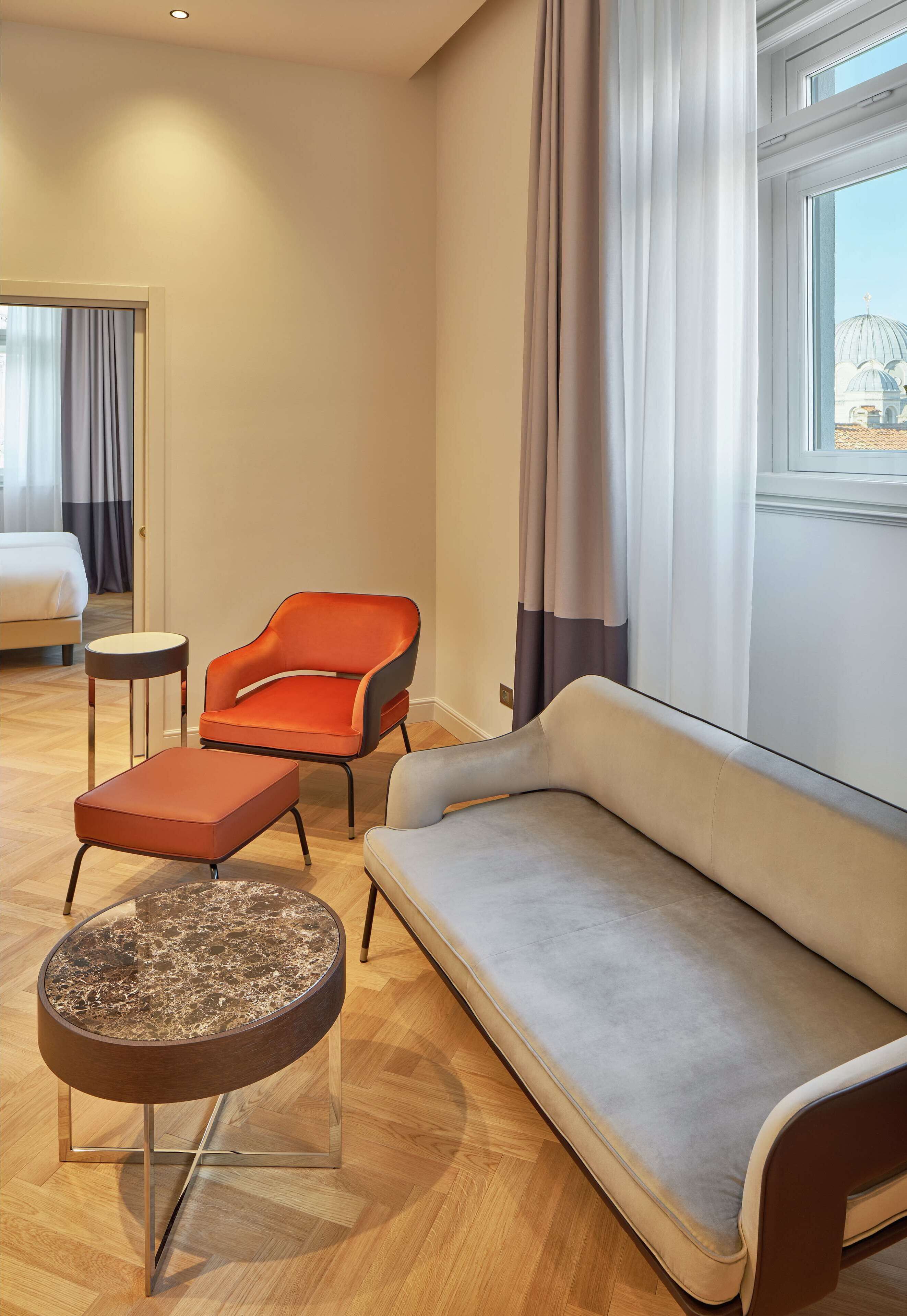 A hotel room with a bed, a grey sofa, an orange armchair, a small round table, and a window with curtains.