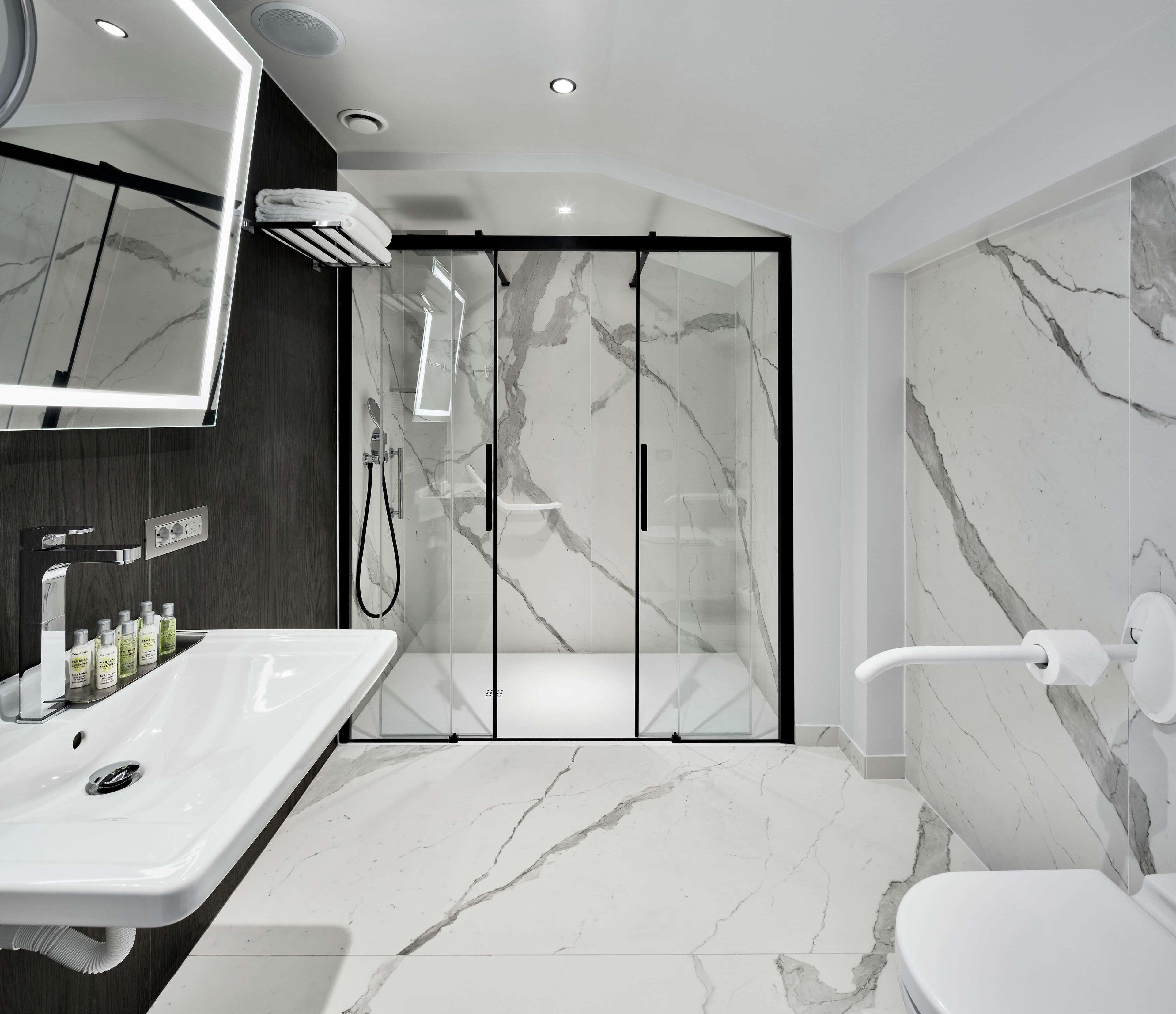 Un baño moderno con un espejo grande, un lavamanos blanco con artículos de aseo, una ducha con mampara de vidrio y suelo de mármol.
