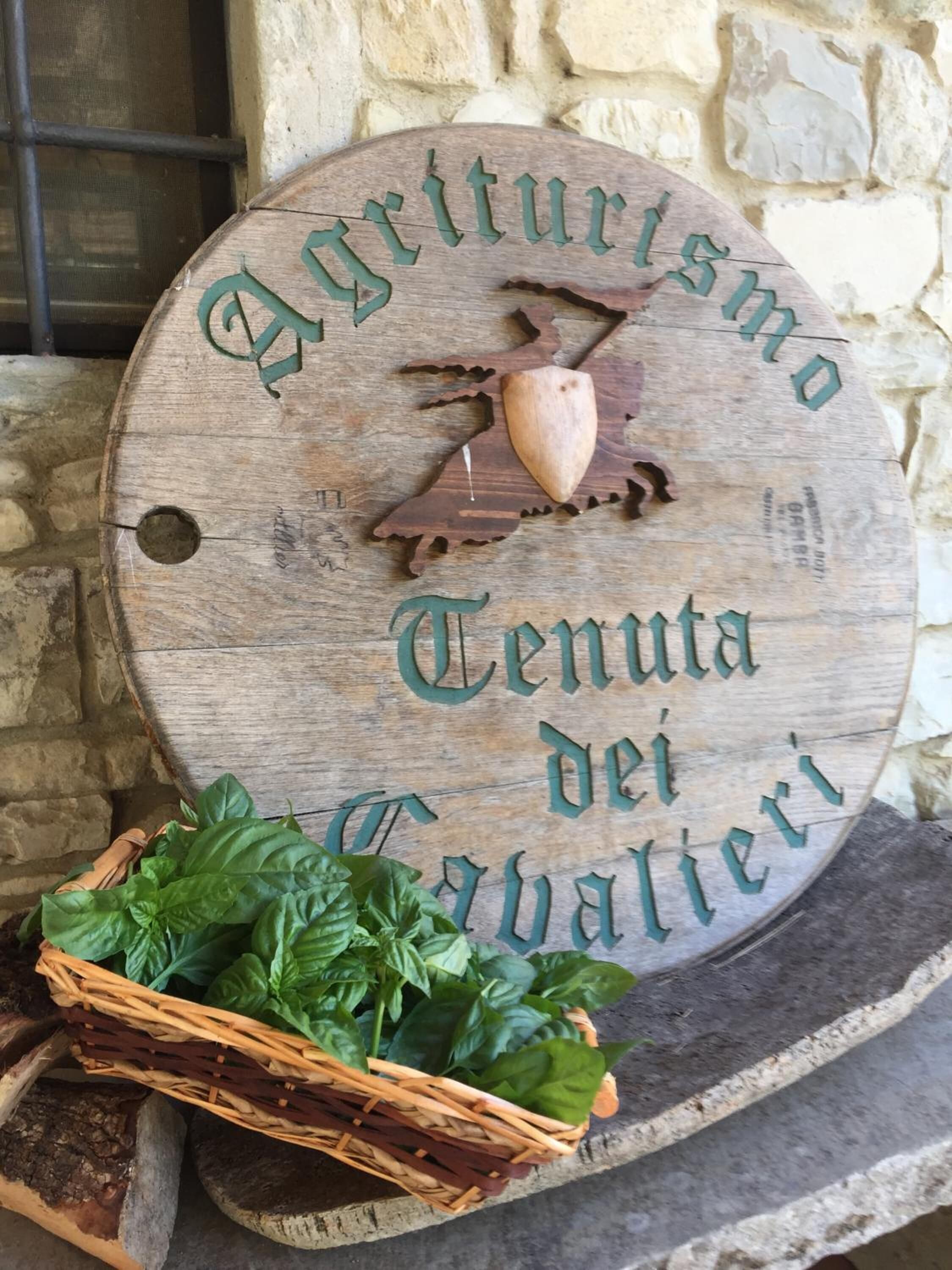 Ein hölzernes Schild mit eingekerbtem Text mit der Aufschrift "Agriturismo Tenuta dei Tabalieri" und einem Wappenschild, an einer Steinsäule befestigt.