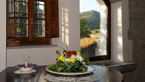Une table ornée d’un centre de table floral, d’une bougie et d’un pichet en verre. Une fenêtre avec des volets en bois offre une vue sur un paysage montagneux.
