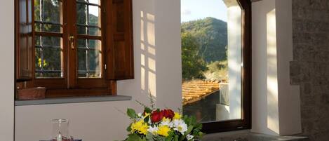 A table with a floral centerpiece, a candle, and a glass pitcher. A window with wooden shutters and a view of a mountain landscape is visible.