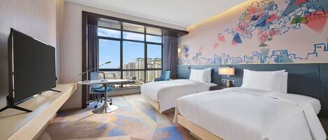A hotel room with two beds, a desk, a TV, and a large window with a city view.