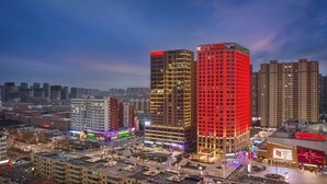 Paisagem urbana noturna com edifícios iluminados, incluindo um hotel com fachada vermelha e um edifício moderno com padrão de grade.