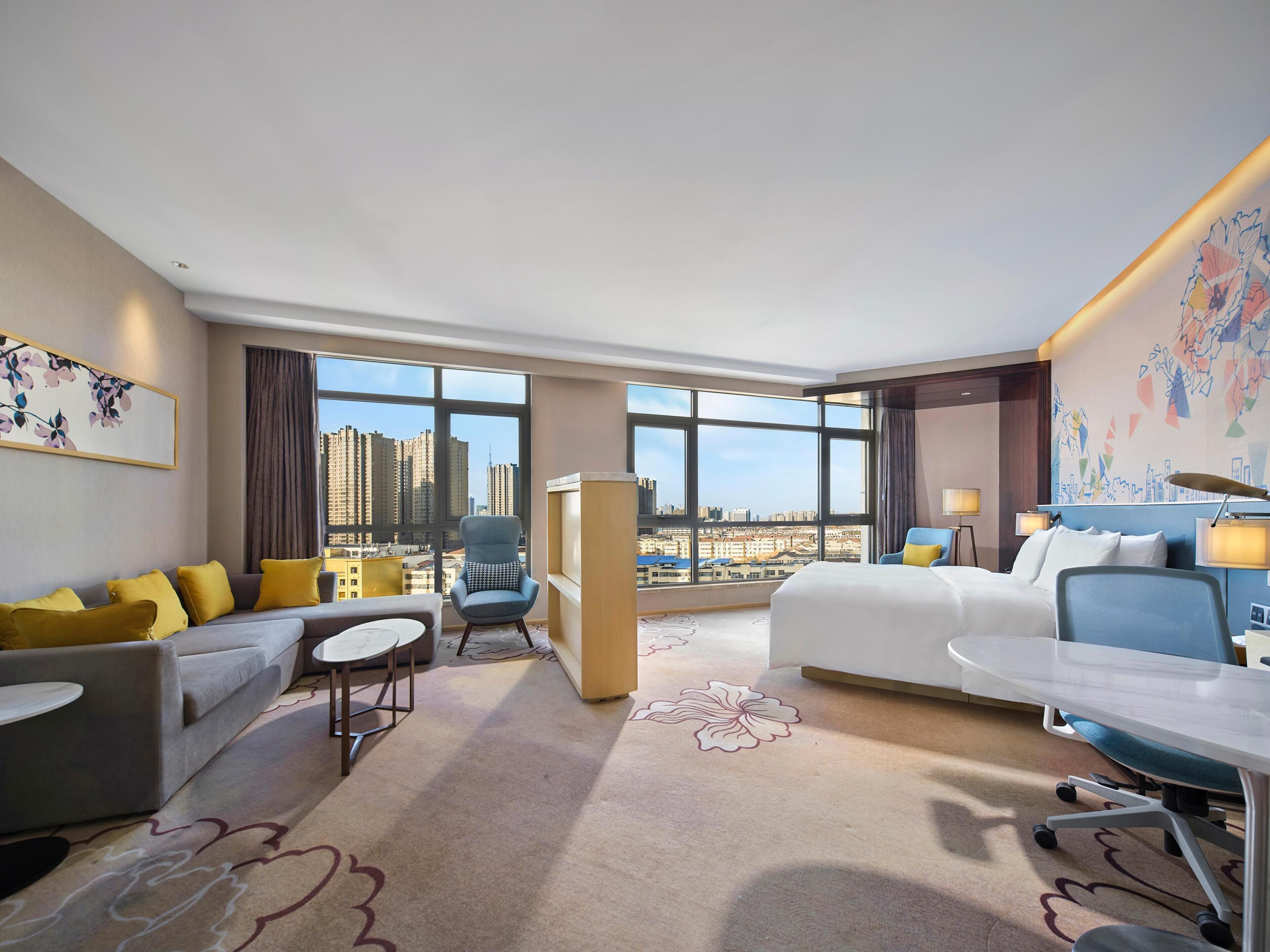 Habitación de hotel con un ventanal grande, una cama, un escritorio, una silla, un sofá y una mesita.