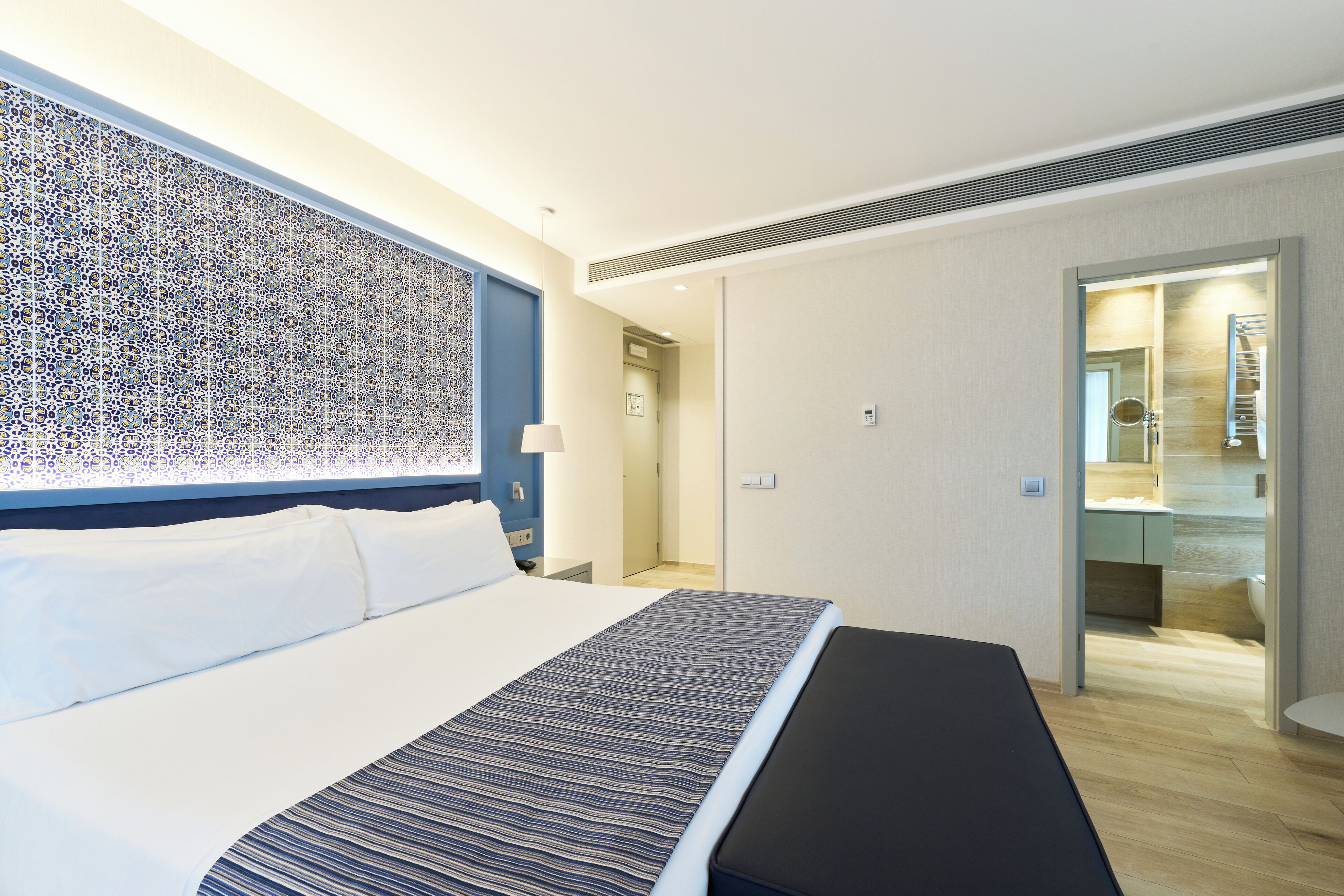 Ein modernes Hotelzimmer mit einem großen Bett, einem gemusterten Kopfteil, einem Nachttisch mit Lampe und Blick ins Badezimmer.