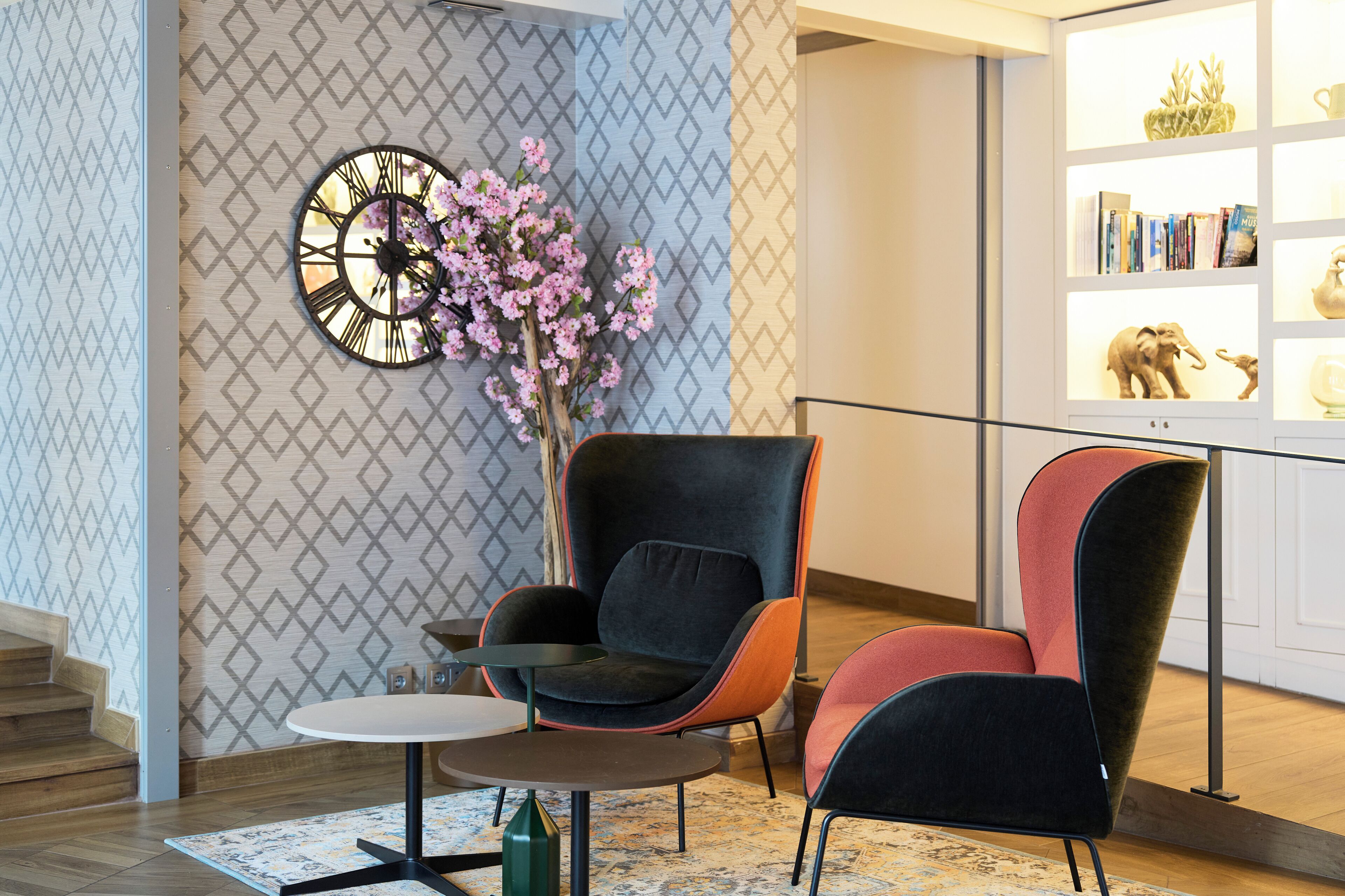 Un intérieur moderne avec un mur orné d’un motif, une horloge, un vase rempli de fleurs roses et une bibliothèque agrémentée d’objets décoratifs.