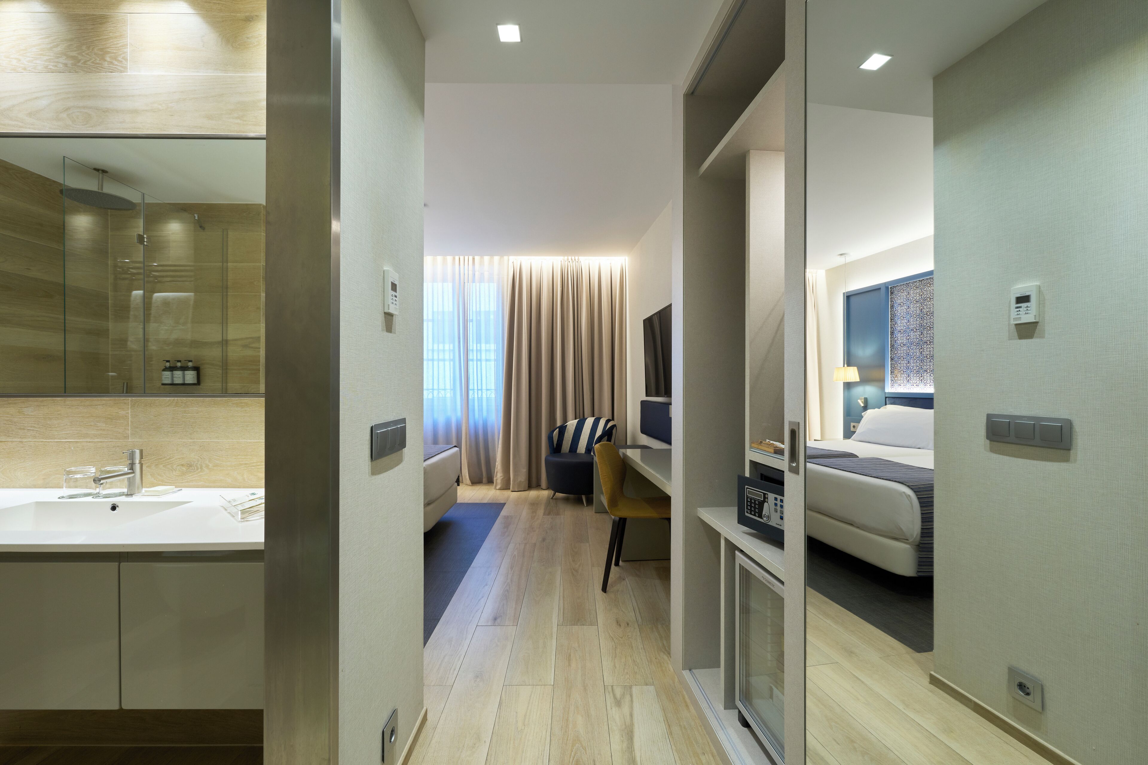 Quarto de hotel moderno com um espelho grande, mesa de cabeceira, cama e banheiro com pia e chuveiro.