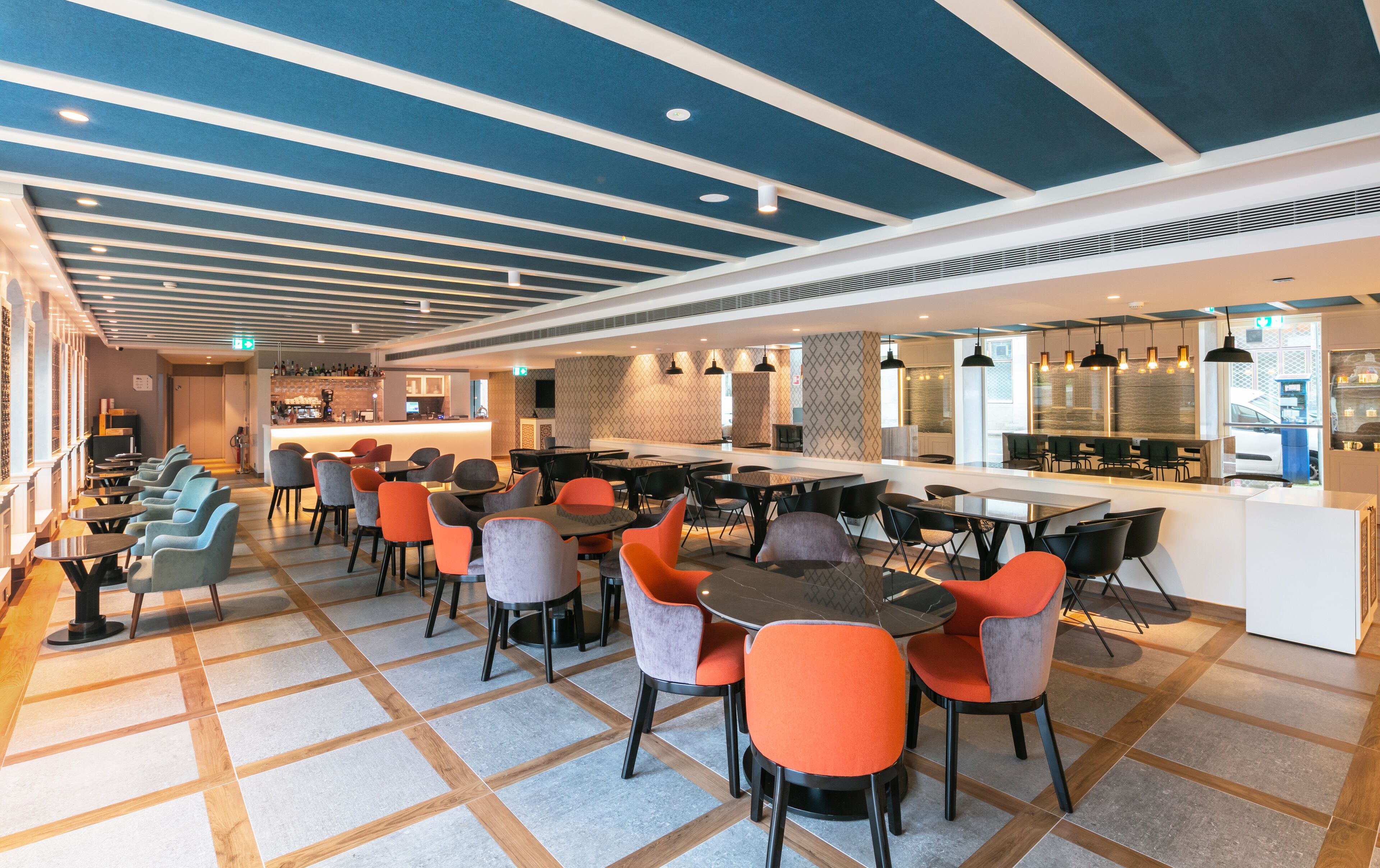 A modern restaurant with a long counter, multiple tables and chairs, and a patterned ceiling.