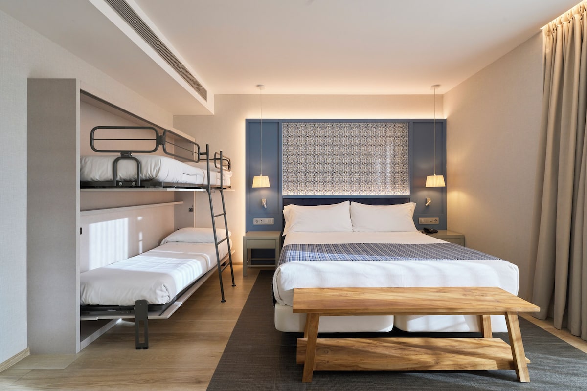 Ein kleines Hotelzimmer mit Etagenbett, einem Einzelbett, einer Holzbänklä und einem gemusterten Kopfteil.