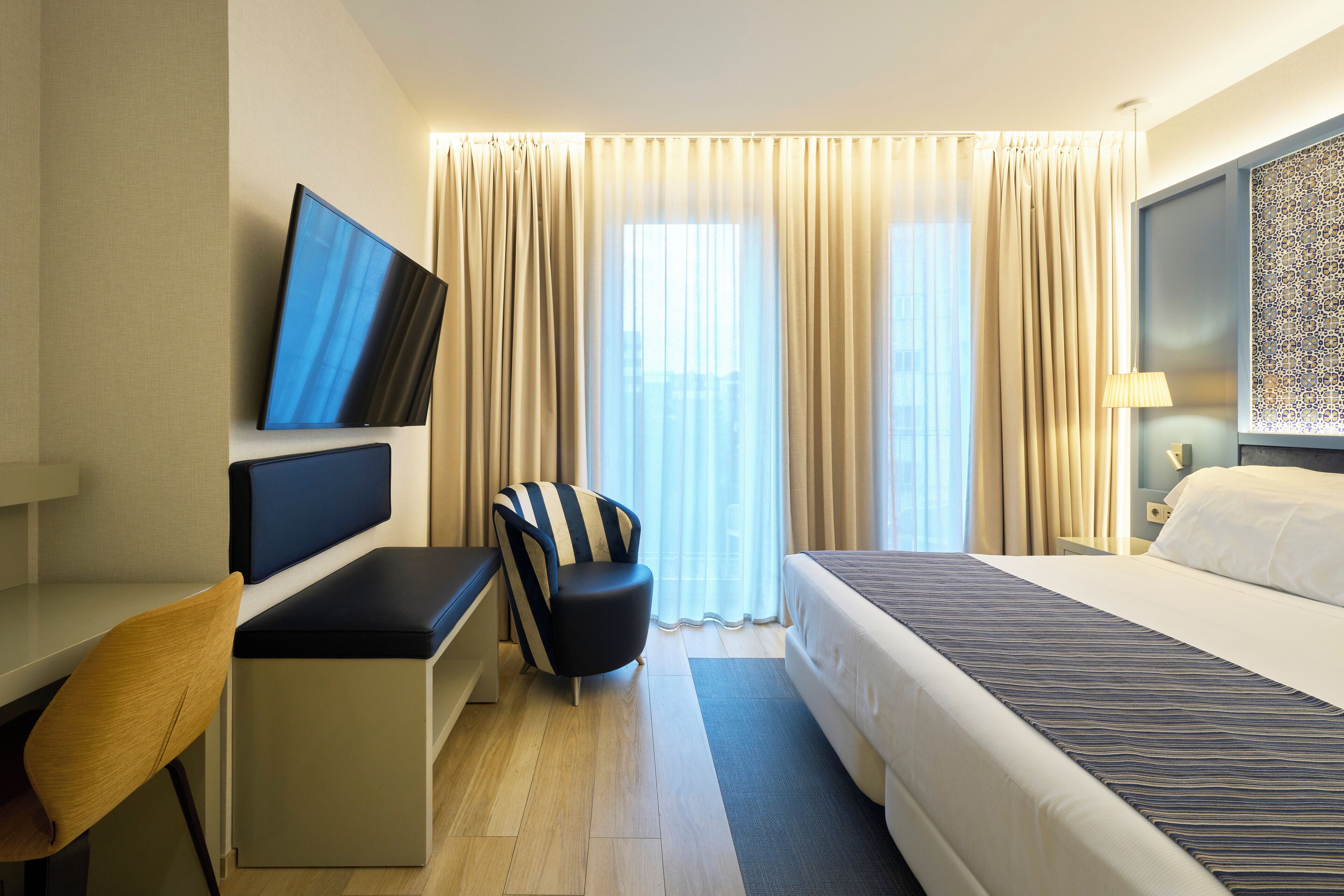 A hotel room with a bed, a desk with a chair, a TV mounted on the wall, and a window with curtains.