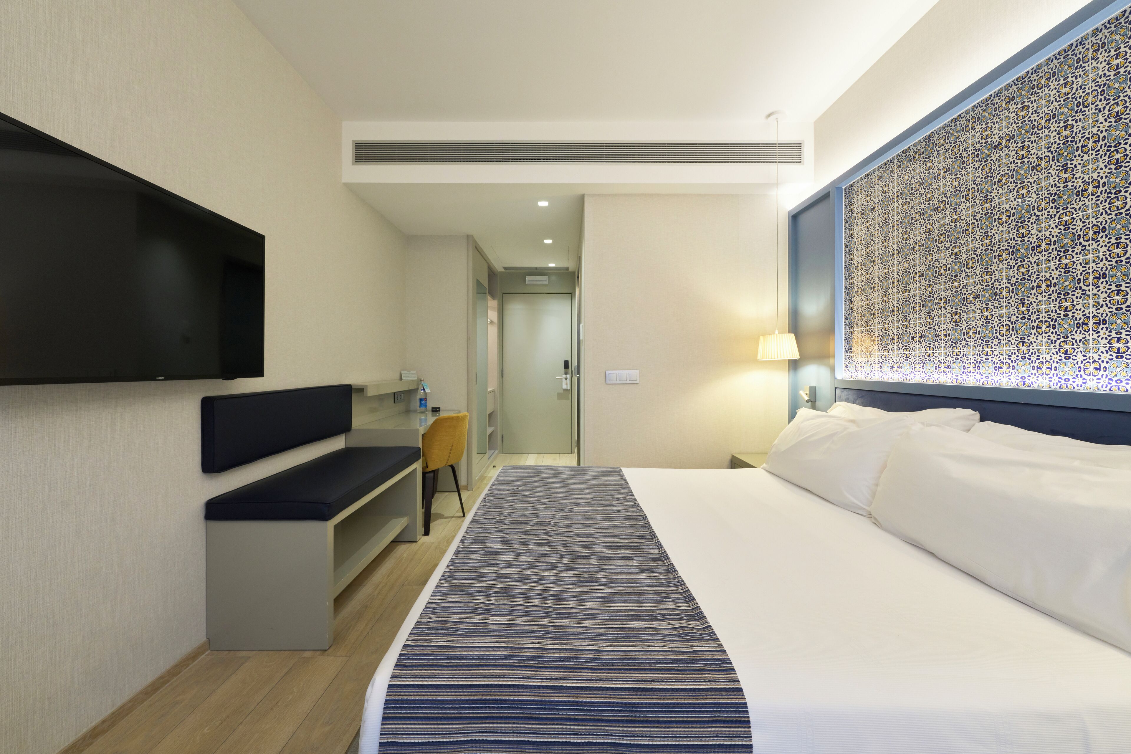 A hotel room with a large bed, a desk with a chair, a TV mounted on the wall, and a decorative wall panel.