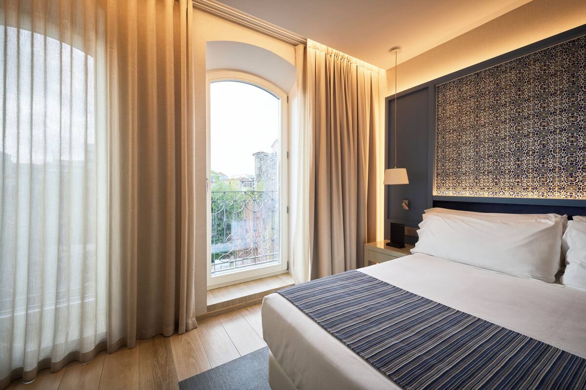 Ein Hotelzimmer mit einem großen Bett, Blick ins Freie durch ein Fenster und einer dekorativen Wandverkleidung.
